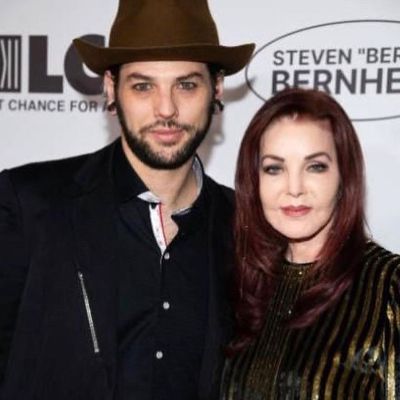 He is wearing all black with brown hat and Priscilla is wearing black and golden stripes dress.